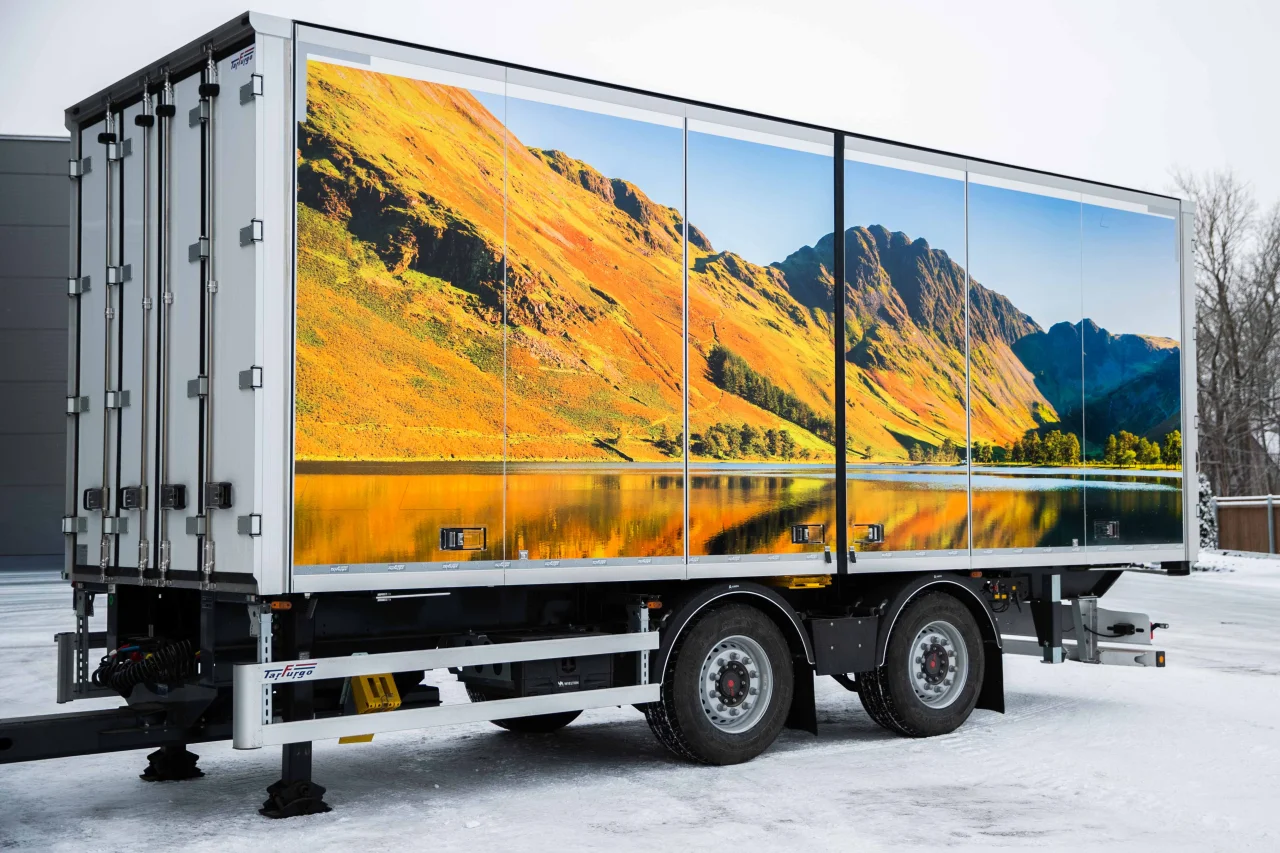 A rear three-quarter view of a white commercial trailer parked in a snowy lot. The trailer is adorned with a vibrant landscape mural showing sun-drenched autumn mountains and a lake. The trailer includes detailed metallic hardware, dual axles, and the "Tesfrigo" logo on the side rail.